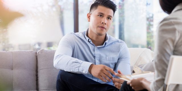 A man with drak brown hair and brown eyes wearing a blue button-down shirt speaks with a therapist.