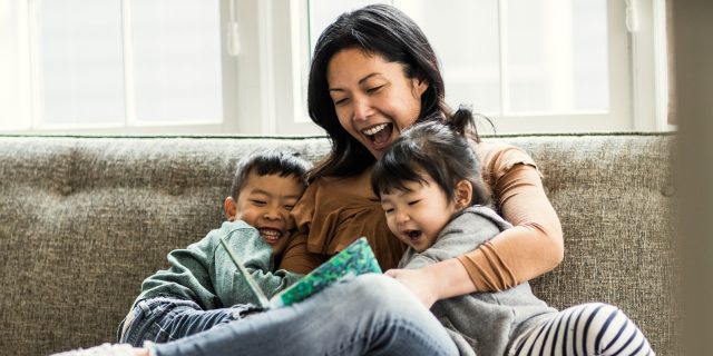 A woman hugs two children, a boy and a girl, as she reads to them. All three are smiling.