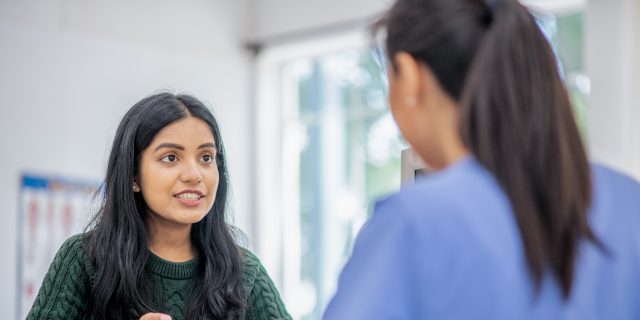 7 Common Stages of Coping With Treatment for a Rare Disease A woman of color wearing a green sweater speaks with a female doctor with long brown hair in a ponytail.