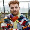 A white man with curly red hair wearing a multicolored striped coat and scarf stands in a garden and looks at the camera.