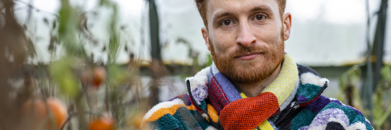 A white man with curly red hair wearing a multicolored striped coat and scarf stands in a garden and looks at the camera.