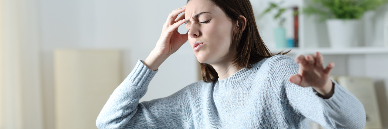 Lessons I Learned From Vertigo and Chronic Illness A white woman with brown hair wearing a sweater hold her head in pain while keeping her other hand extended for balance.