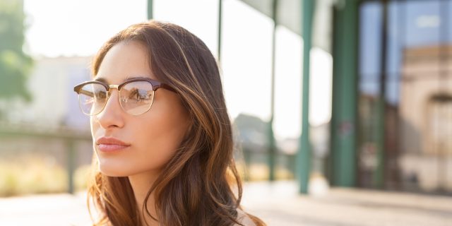 A woman with long brown hair and glasses wearing an off-shoulder shirt stares into the distance outside.