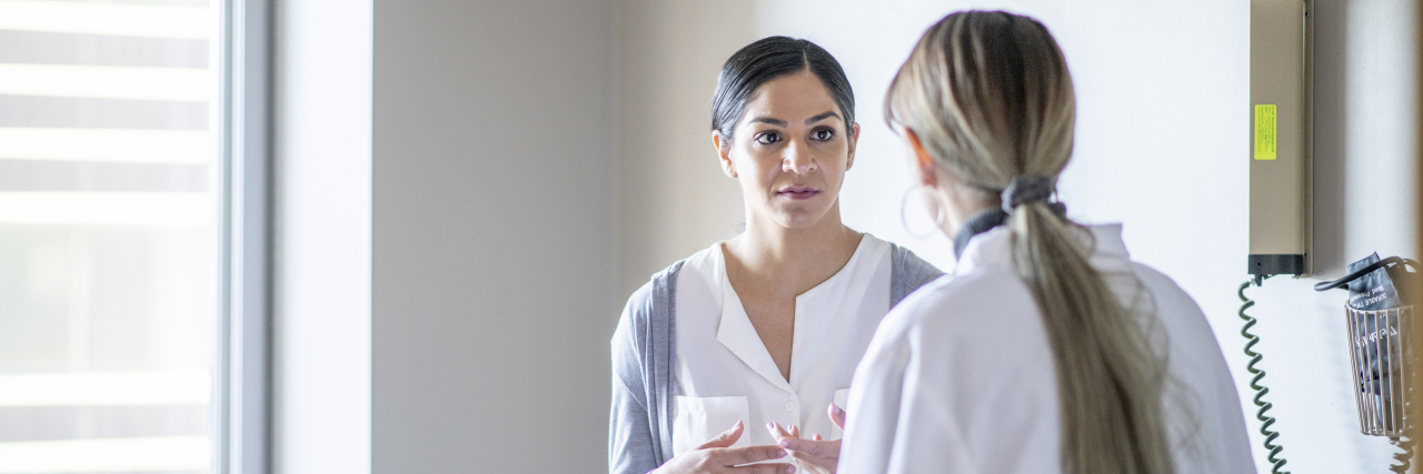 Long Wait Times for Doctor's Appointments Harm Patients A woman of color wearing a white blouse and gray cardigan sits on an exam table while speaking with a female doctor with long blonde hair.