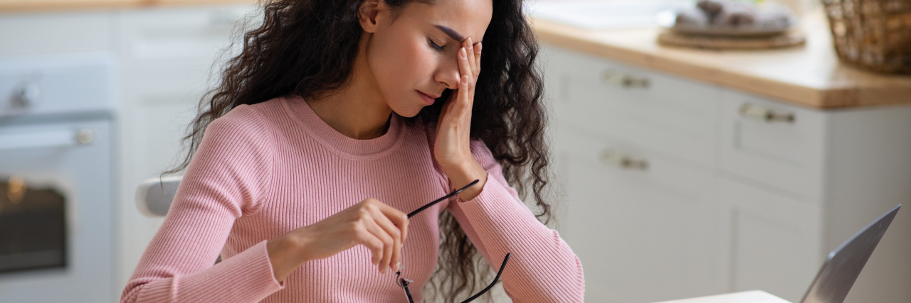 Imagine My Daily Life as a Chronically Ill Person A woman with curly black hair wearing a pink shirt holds her glasses while hunched over a laptop in pain.