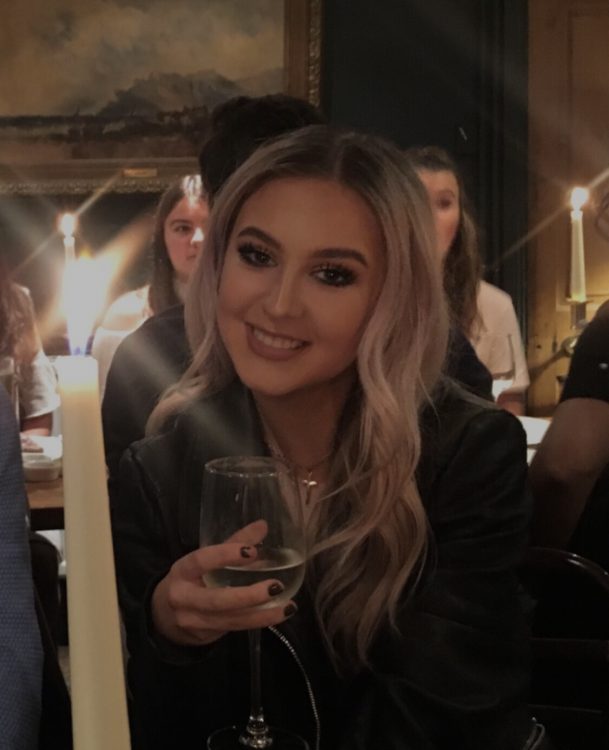 A white woman with wavy blonde hair wearing a black blazer smiles while holding a glass.