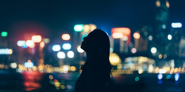 Silhouette of woman looking up towards sky against illuminated and multi-colored cityscape at night
