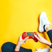 Young thin woman playing with red gamepad, sitting on yellow background. flat lay.