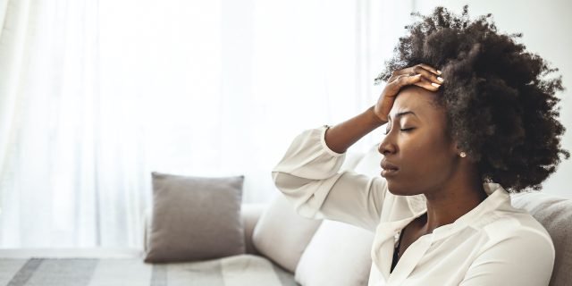 Black woman holding her hand on her head, sitting on the couch with her eyes closed