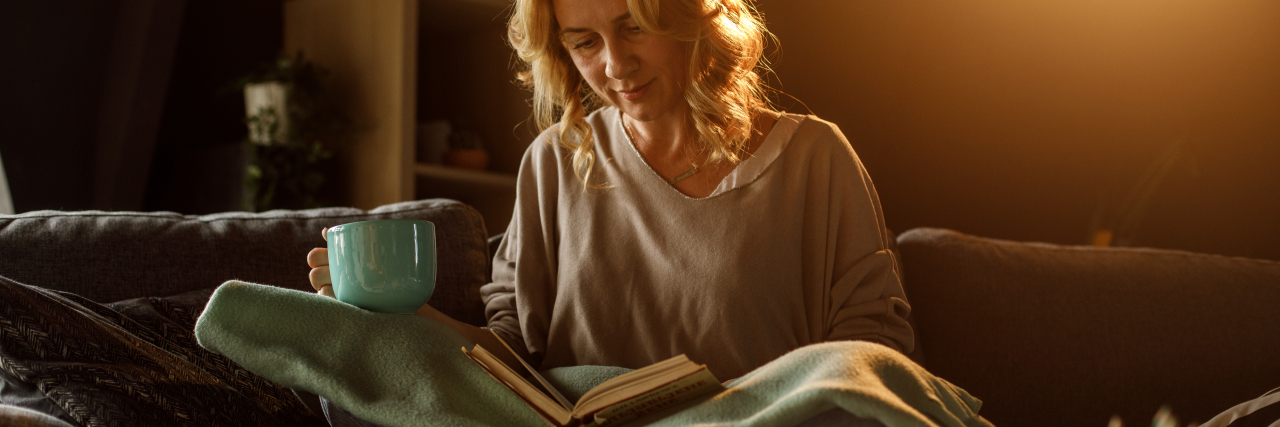8 Books About Migraine You'll Want to Read Books about migraine. Woman reading a book and enjoying tea on the sofa..