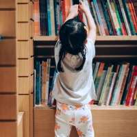 Young Asian girl reaching up to get books from bookshelf in a library