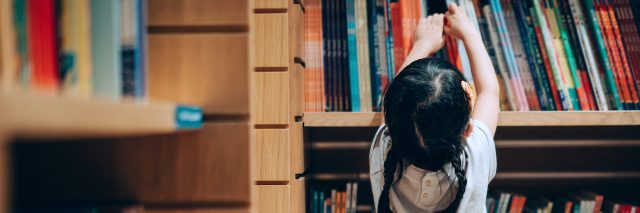 Young Asian girl reaching up to get books from bookshelf in a library