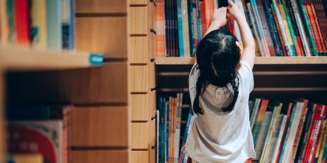 12 Children's Books That Smash the Patriarchy Young Asian girl reaching up to get books from bookshelf in a library