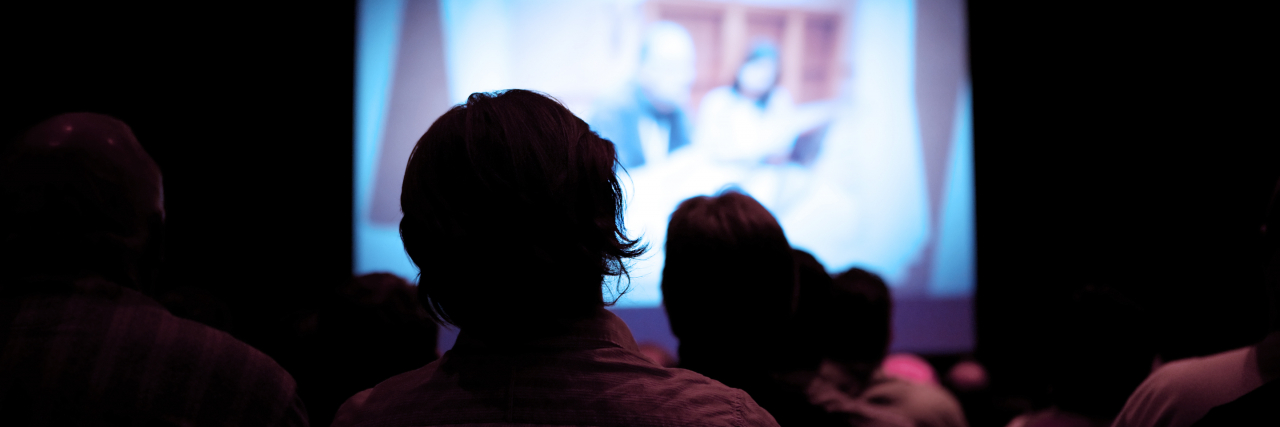 3 Tips for Safely Enjoying Triggering Media An unknown number of people sitting in a dark theater. There is a projector screen in front of them with a man and woman at a conference.