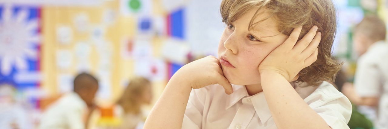 My School Reports Show My Undiagnosed Childhood ADHD photo of a young boy in school with his head on his hands, looking distracted while other children work