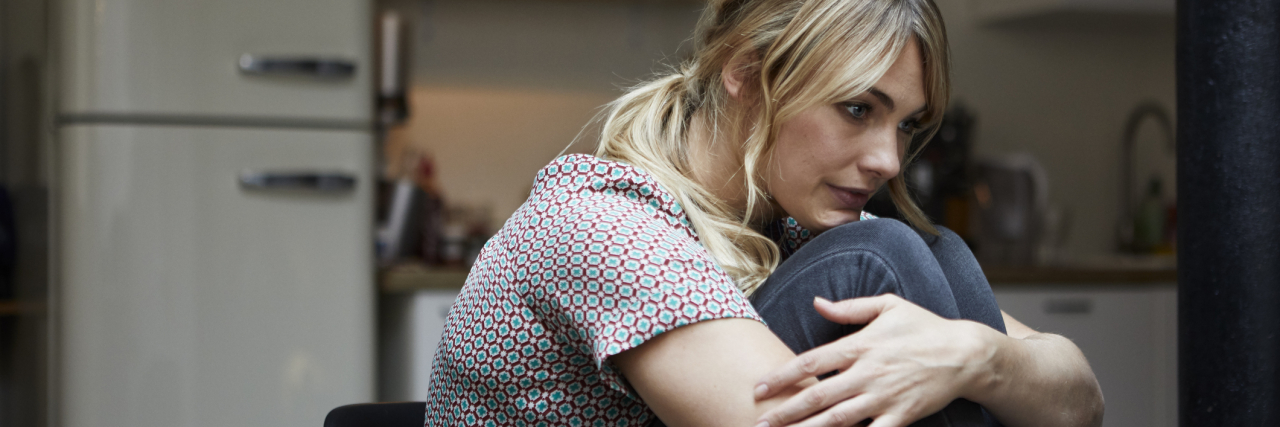 photo of a young woman sitting at a table, hugging her legs and looking sad or lonely