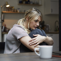 photo of a young woman sitting at a table, hugging her legs and looking sad or lonely