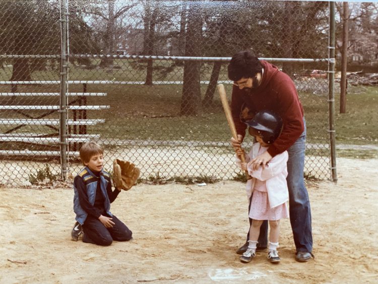 Photo of Francesca as a young girl holding a baseball bat with her Dad, her brother playing catcher behind her