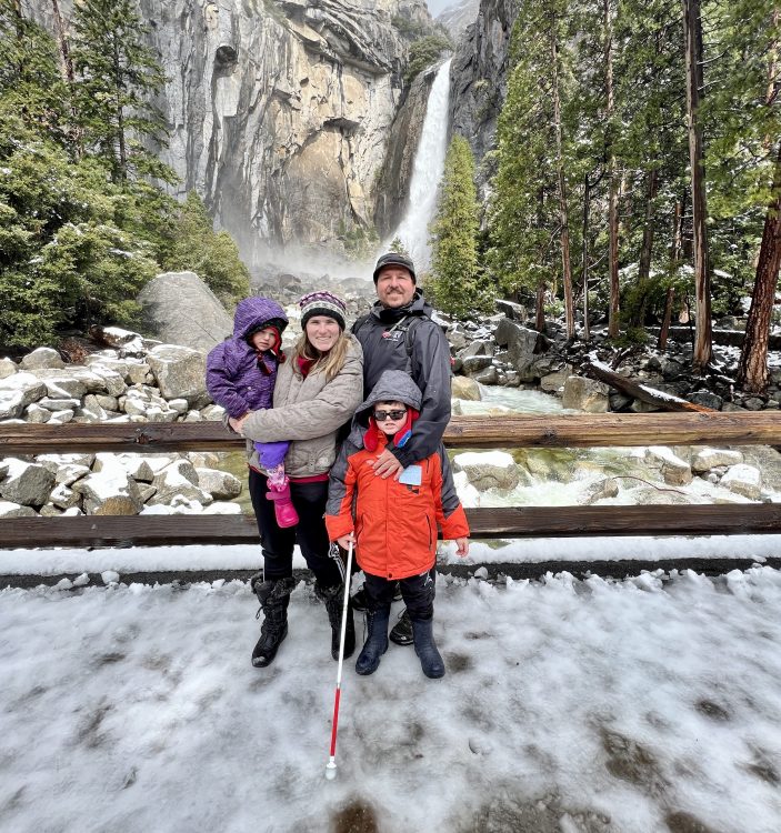 Karl standing on snow-covered ground with his cane, in front of waterfall at Yosemite on a hike with his family (mother, father and sister)