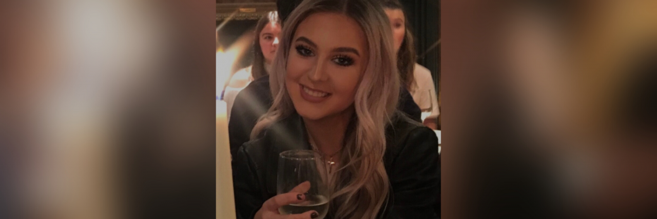 A white woman with wavy blonde hair wearing a black blazer smiles while holding a glass.