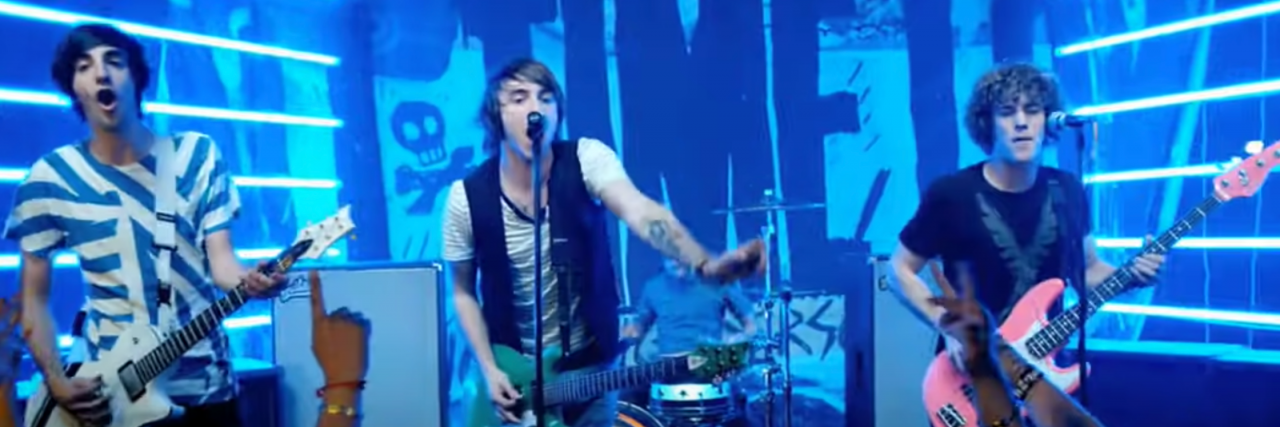 What 8 Pop Punk Bands Tell Us About Mental Health in Your Teen Years A screenshot of All Time Low's music video for "Weightless." The boys stand on stage mid song with the band's banner behind them with the band's name in bold lettering.