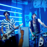 A screenshot of All Time Low's music video for "Weightless." The boys stand on stage mid song with the band's banner behind them with the band's name in bold lettering.