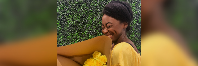A very pretty Black girl glowing in yellow stands in front of a grass wall with a big bouquet of yellow roses. She's smiling wide. She's really pretty. Yes she's me.