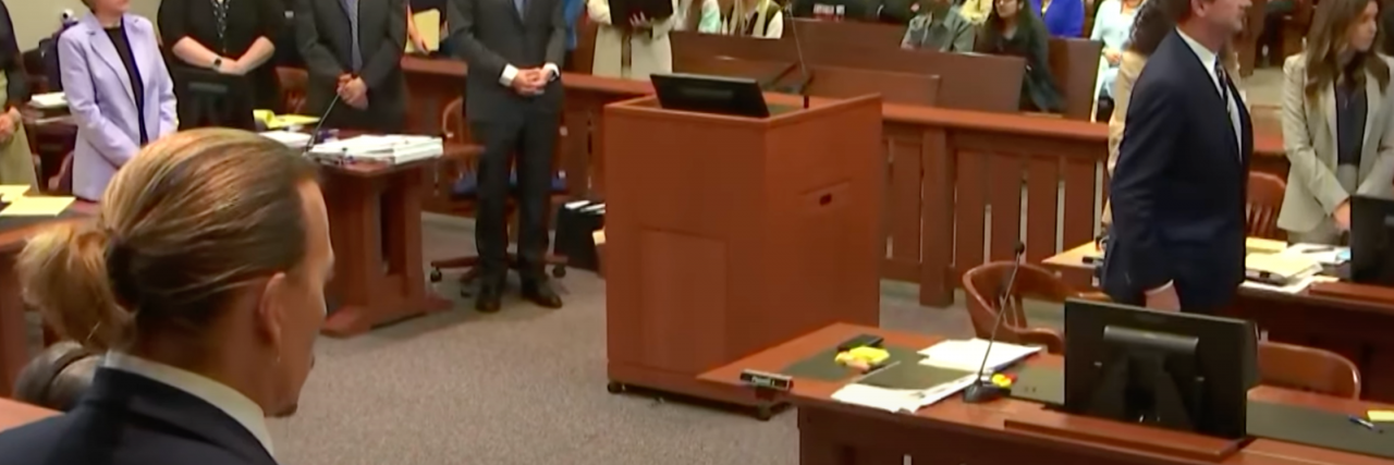A screencap from an Entertainment Tonight video of Johnny Depp at trialk. They're in a courtroom with different people in business clothing standing around at the beginning of the proceedings.