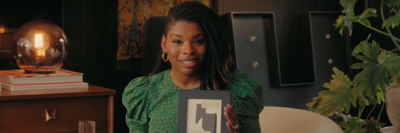A screen cap of Carmeon Hamilton wearing a beautiful green shirt holding up some homemade art. They have dreadlocks and are sitting in a living room.