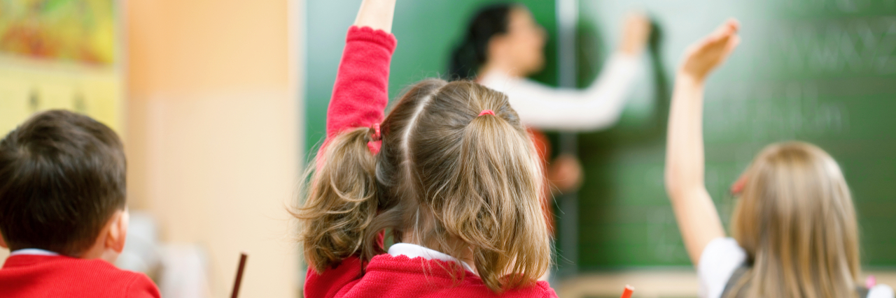 What Has Helped Me and Other Students with Visual Processing Disorder A young girl wearing a red sweater raises her hand in a classroom while surrounded by her classmates.