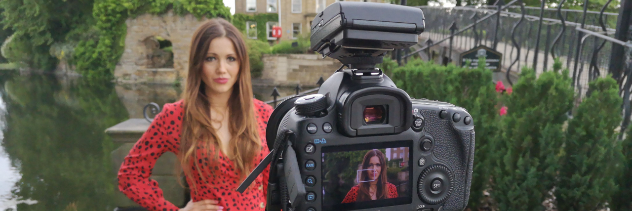 Why I'm Talking About Incontinence and Anxiety Contributor wearing a red dress and standing by a picturesque river and bridge, with a SLR camera on a tripod pointed at her