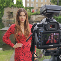Contributor wearing a red dress and standing by a picturesque river and bridge, with a SLR camera on a tripod pointed at her