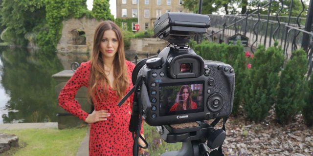 Contributor wearing a red dress and standing by a picturesque river and bridge, with a SLR camera on a tripod pointed at her