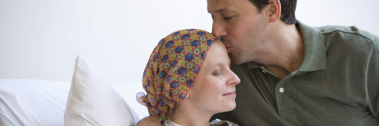 A man with brown hair kisses a woman's forehead in the hospital. She is wearing a hospital gown and headscarf.