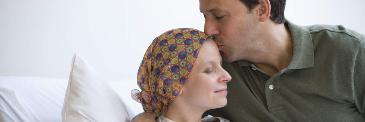 How You Can Really Help Cancer Patients' Caregivers A man with brown hair kisses a woman's forehead in the hospital. She is wearing a hospital gown and headscarf.