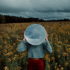 Person standing in field of yellow flowers, holding a white round mirror which reflects the cloudy sky