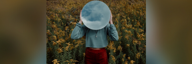 Person standing in field of yellow flowers, holding a white round mirror which reflects the cloudy sky