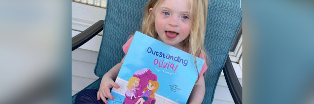 Olivia, a little girl with Down syndrome holding the children's book about her, "Outstanding Olivia."