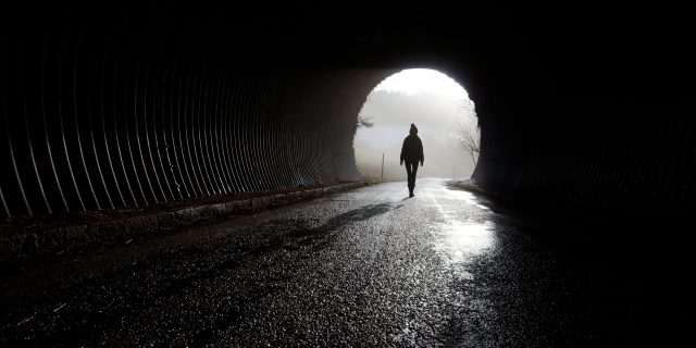 A silhouette of a person walking through a dark tunnel towards a patch of light.