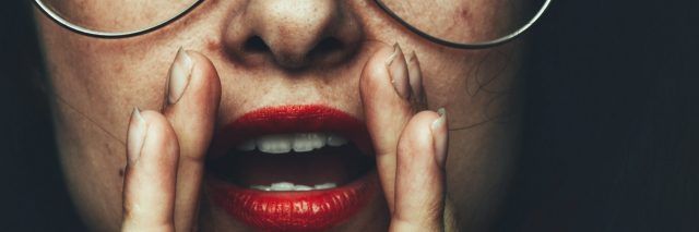 A person with freckles and red lipstick puts their hands around their mouth.