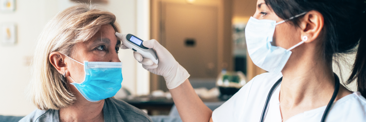 Feeling Exhausted After Fighting COVID-19 for Weeks A woman with blonde hair gets her temperature taken by a female doctor with black hair. Both are wearing face masks.