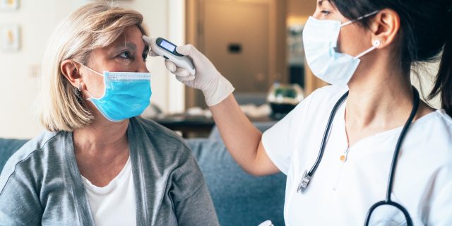 A woman with blonde hair gets her temperature taken by a female doctor with black hair. Both are wearing face masks.