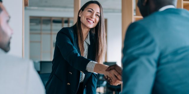 A woman with long, brown hair wearing a green blazer and a button-down shirt shakes the hand of a black man.