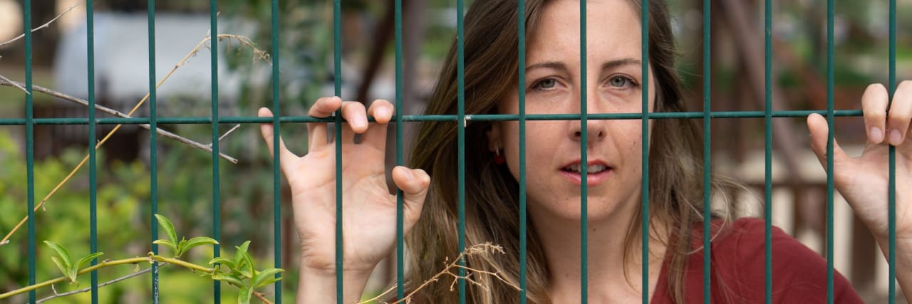A brunette woman with blue eyes and a red shirt holds onto a green fence and pokes her fingers through the gaps as if to escape.