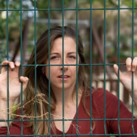 A brunette woman with blue eyes and a red shirt holds onto a green fence and pokes her fingers through the gaps as if to escape.