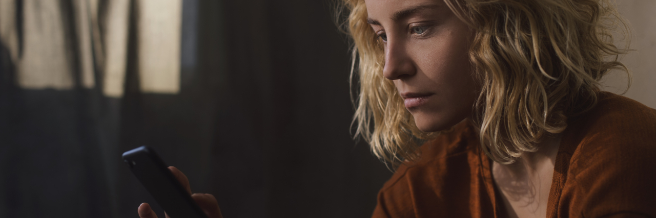 Letting Go of a Deceased Child and Feeling Betrayal A woman with wavy blonde hair and blue eyes wearing an orange top looks pensively at her cell phone.