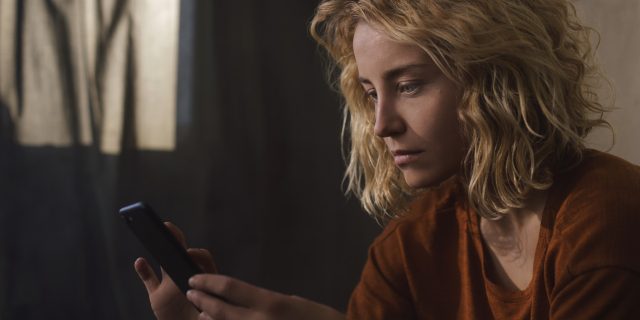 A woman with wavy blonde hair and blue eyes wearing an orange top looks pensively at her cell phone.