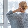A blonde woman wearing a gray sweater scratches her head as she looks out the window..