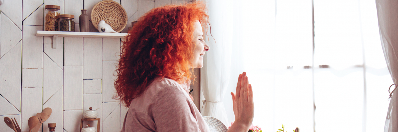 A woman with curly red hair wearing a pink top waves out a window while standing in a kitchen.