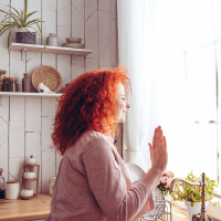 A woman with curly red hair wearing a pink top waves out a window while standing in a kitchen.